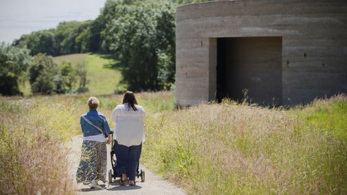 Two adults walk along a narrow path through tall grass, pushing a stroller. To the right stands a round concrete architectural artwork structure with a large rectangular opening. Rolling countryside and trees extend into the distance under a bright sky.